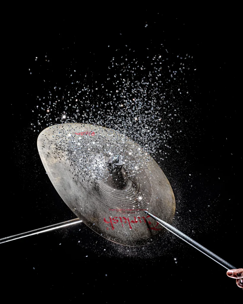 Cymbal struck in a burst of silver powder, frozen mid-strike against black