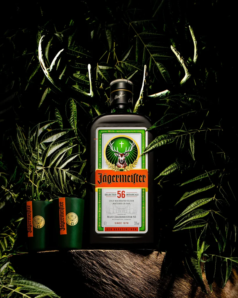 Herbal liqueur bottle and two shot glasses among dark foliage and antlers (retouched)