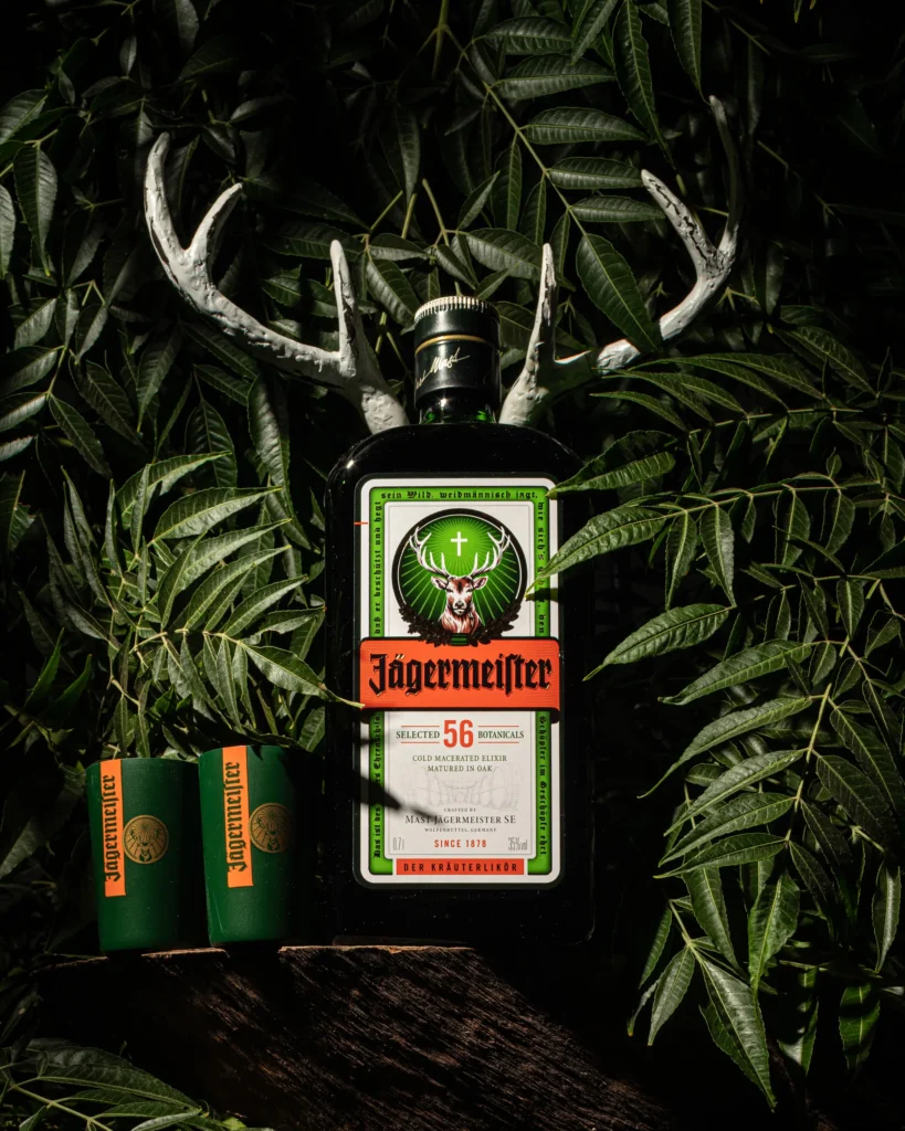 Herbal liqueur bottle and two shot glasses among dark foliage and antlers (unretouched original)